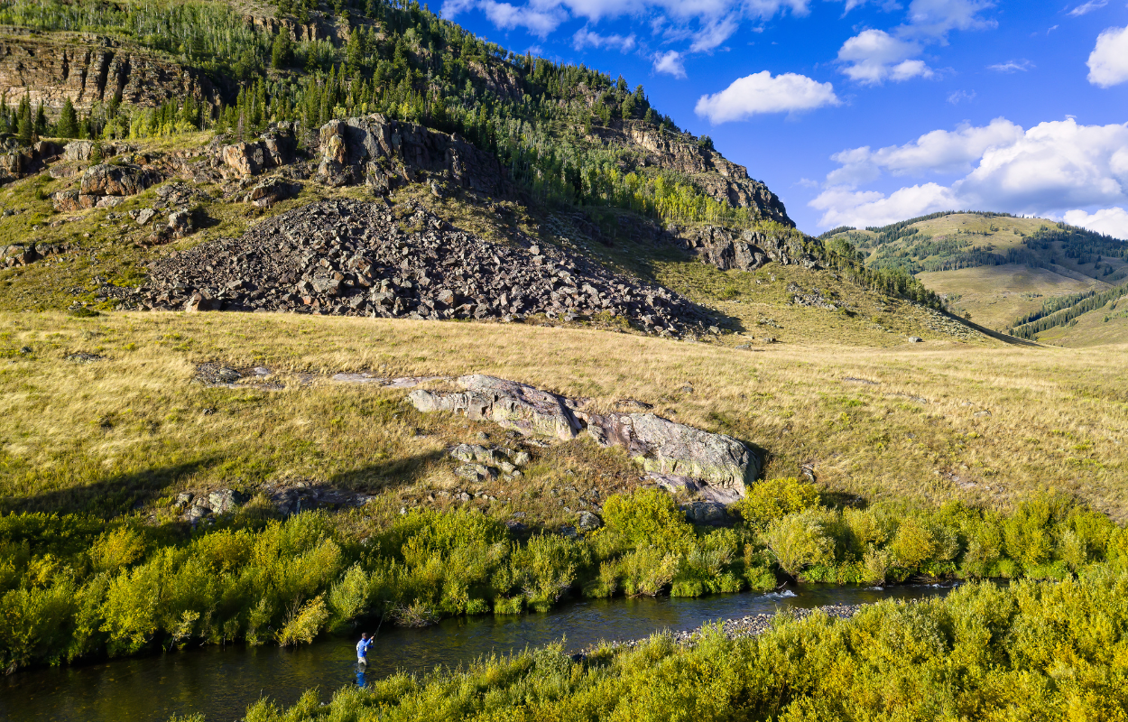 an angler stands in a small stream while fly fishing for trout in the mountains